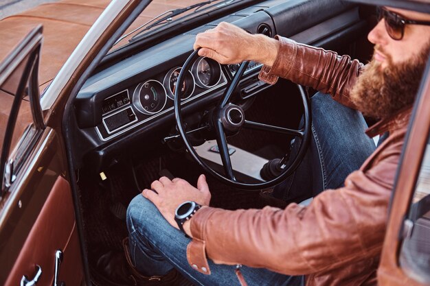 Stylish bearded male in sunglasses dressed in brown leather jacket sits behind the wheel of a tuned retro car with open door.