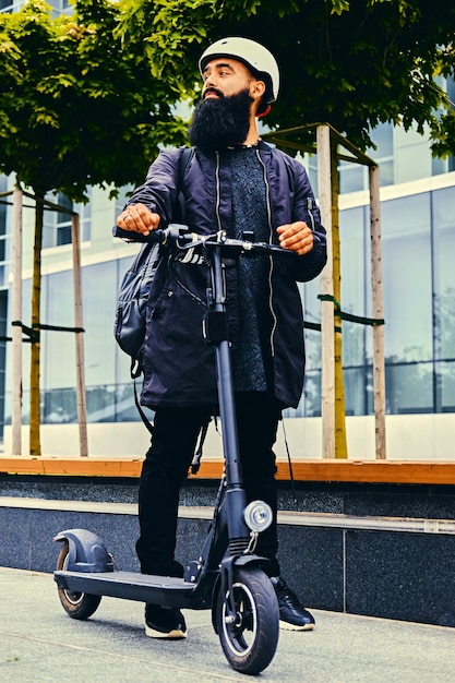 Stylish bearded male in sunglasses posing on electric scooter in over modern building background.