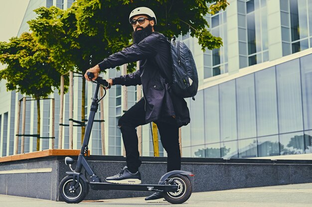 Stylish bearded male in sunglasses posing on electric scooter in over modern building background.