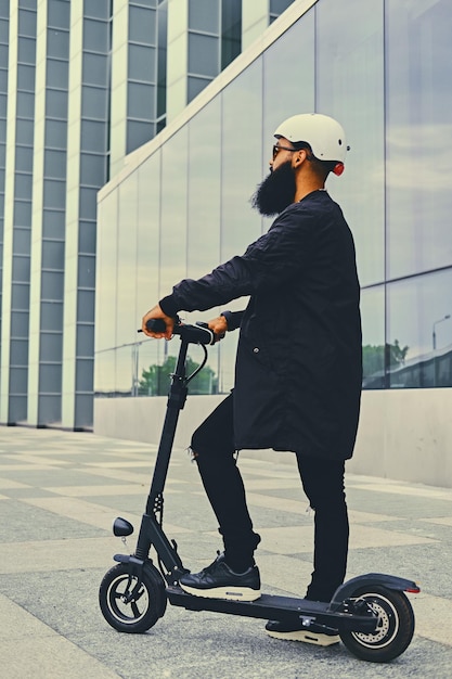 Stylish bearded male in sunglasses posing on electric scooter in over modern building background.