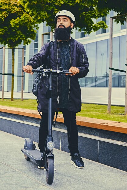 Stylish bearded male in sunglasses posing on electric scooter in over modern building background.