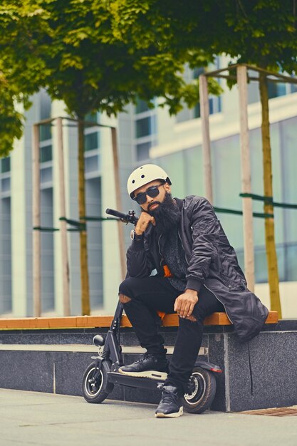 Stylish bearded male in sunglasses relaxing on a bench after riding by electric scooter in downtown.