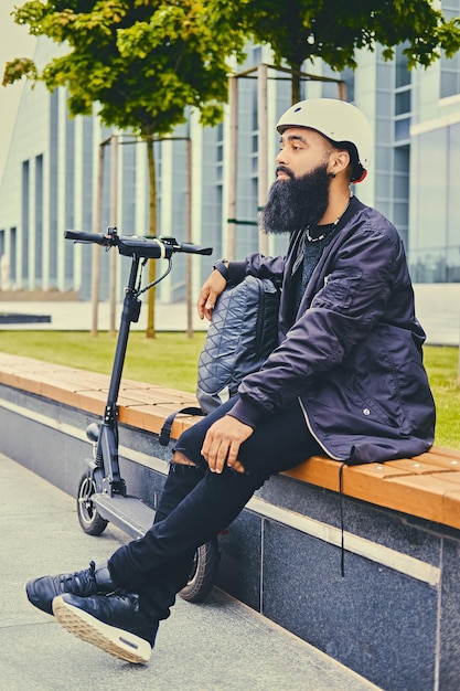 Free photo stylish bearded male in sunglasses relaxing on a bench after riding by electric scooter in downtown.