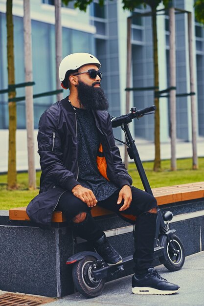 Stylish bearded male in sunglasses relaxing on a bench after riding by electric scooter in downtown.