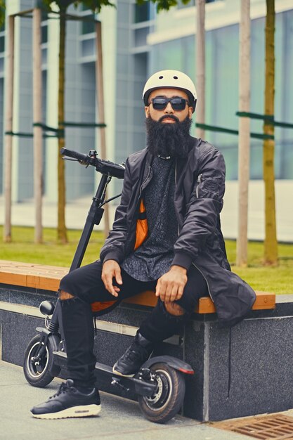 Stylish bearded male in sunglasses relaxing on a bench after riding by electric scooter in downtown.