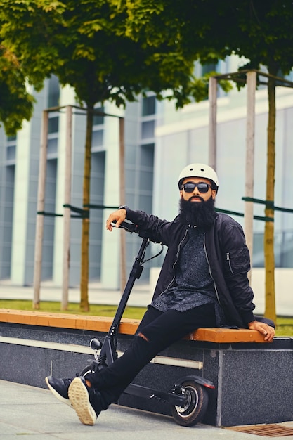 Free photo stylish bearded male in sunglasses relaxing on a bench after riding by electric scooter in downtown.