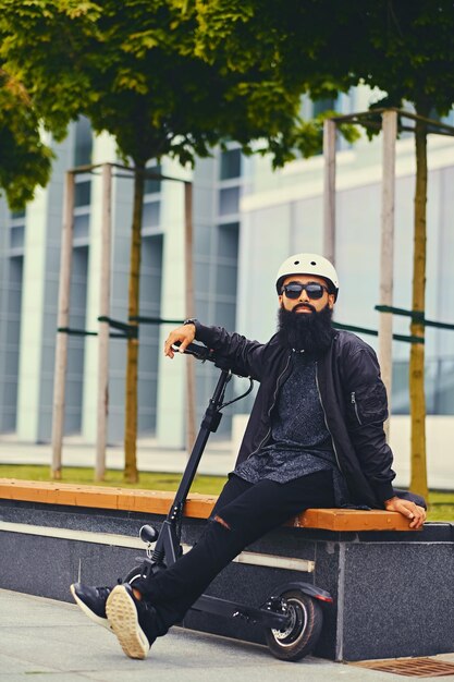 Stylish bearded male in sunglasses relaxing on a bench after riding by electric scooter in downtown.