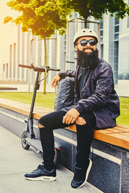 Стильный бородатый мужчина в солнцезащитных очках отдыхает на скамейке после катания на электрическом скутере в центре города.