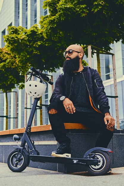 Stylish bearded male in sunglasses relaxing on a bench after riding by electric scooter in downtown.