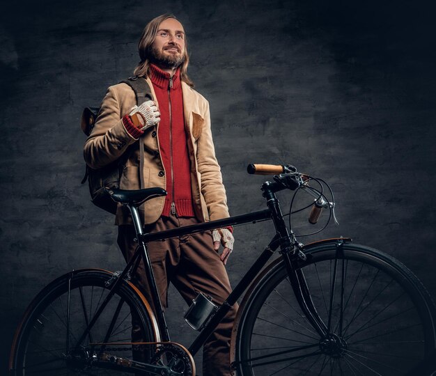 Стильный бородатый мужчина с длинными волосами держит рюкзак и позирует возле фиксированного велосипеда в студии.