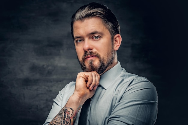 Stylish bearded male with tattooed arms, dressed in a shirt posing over grey background.