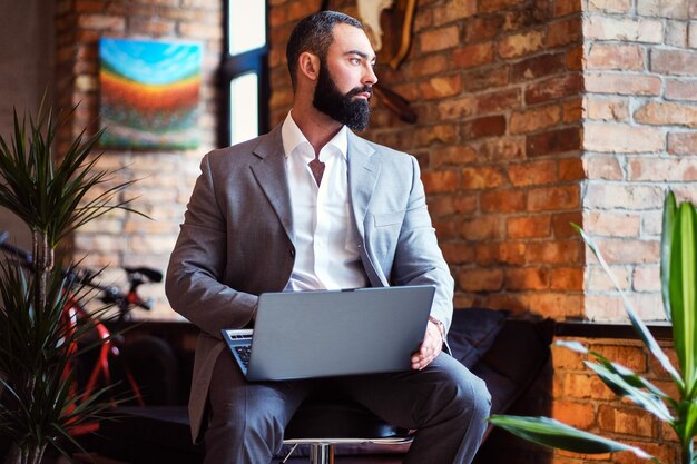 Стильный бородатый мужчина работает с ноутбуком в комнате с лофтовым интерьером.