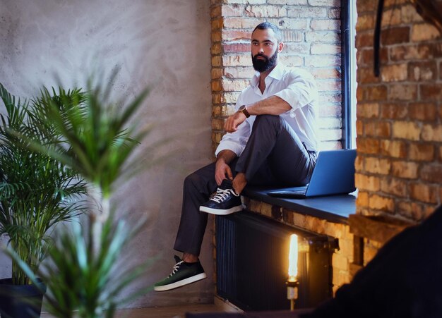 Stylish bearded male works with a laptop in a room with loft interior.