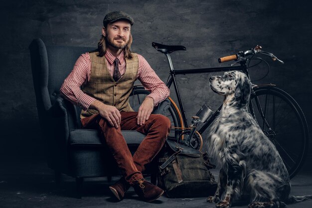 A stylish bearded man in a cap sits on a chair with an Ireland setter dog and a bicycle on a background.