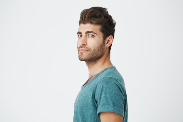 Stylish bearded man with appealing dark eyes seriously . Handsome hipster guy with trendy hairstyle and beard on white wall