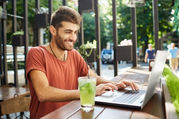 Стильный бородатый мужчина с темными волосами, широко улыбающийся, в красной футболке, занят работой, пьет лимонад, освежая себя лимонадом. Фрилансер, работающий на открытом воздухе, сидя за деревянным столом с не