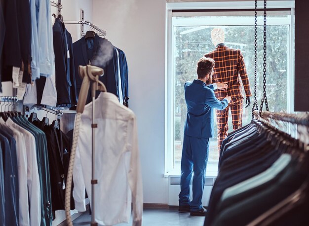 Stylish bearded seller care about suit on a mannequin in a menswear store.