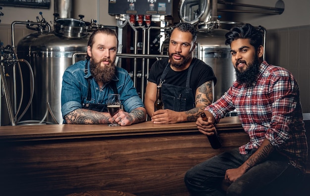 Stylish bearded tattooed Caucasian, Black and Indian men drinking craft beer in the microbrewery.