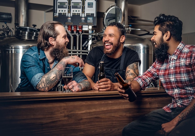 Стильные бородатые татуированные белые, черные и индийские мужчины пьют крафтовое пиво в пивоварне.