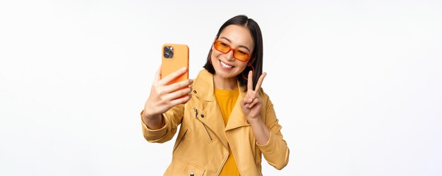 Stylish beautiful asian girl in sunglasses taking selfie on smartphone posing for photo holding mobile phone white studio background