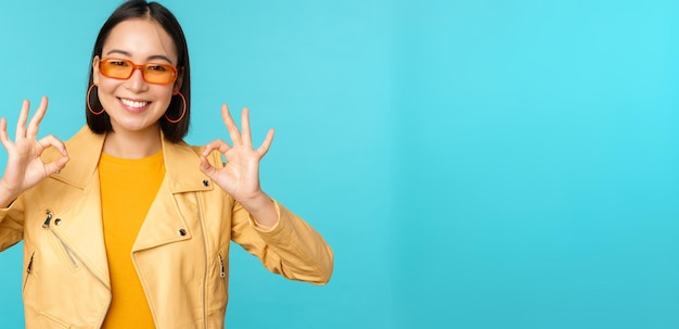 Stylish beautiful asian woman in sunglasses smiling amazed showing okay ok sign recommending smth supports excellent choice standing over blue background