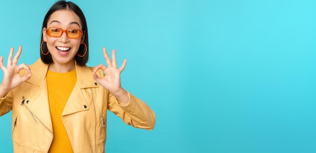 Stylish beautiful asian woman in sunglasses smiling amazed showing okay ok sign recommending smth supports excellent choice standing over blue background