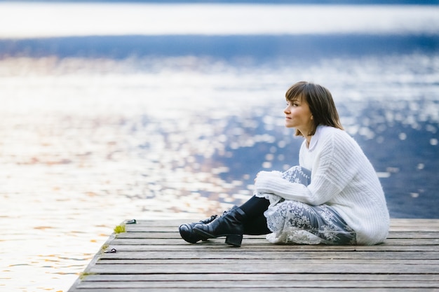 A stylish and beautiful girl sits on a bridge near a large lake