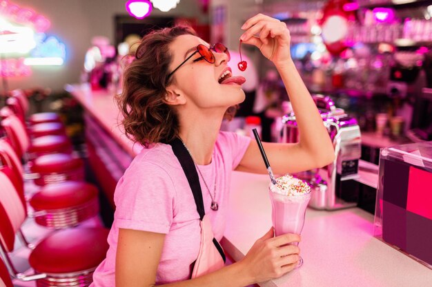 복고풍 빈티지 50년대 카페에 있는 세련된 아름다운 섹시한 여성이 분홍색 티셔츠에 밀크 쉐이크 칵테일을 마시는 바에 앉아 체리를 먹으면서 즐거운 시간을 보내고 있습니다.