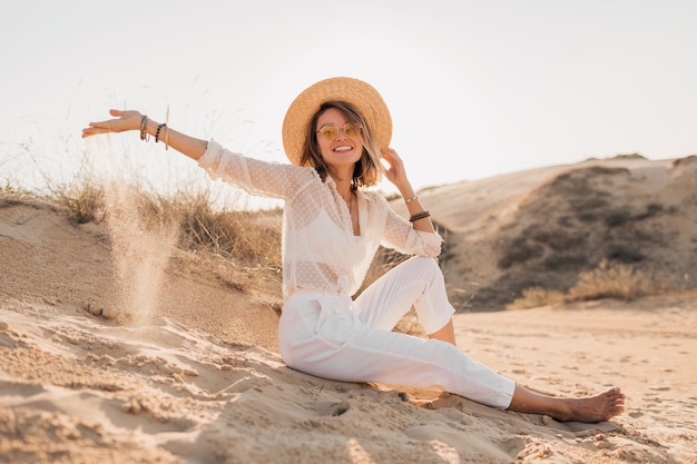 Stylish beautiful woman in desert sand in white outfit wearing straw hat on sunset