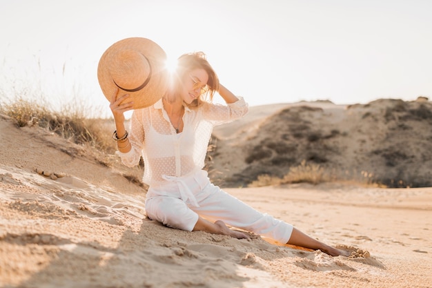 Stylish beautiful woman in desert sand in white outfit wearing straw hat on sunset
