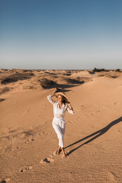 Stylish beautiful woman walking in desert sand dressed in white trousers and blouse wearing straw hat on sunset