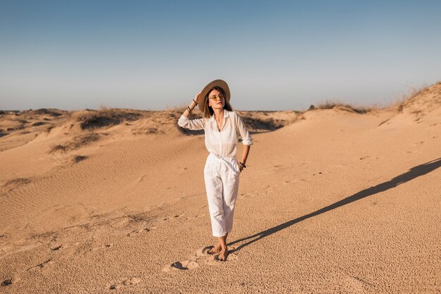 일몰에 밀짚 모자를 쓰고 흰 옷을 입고 사막 모래에서 걷는 세련된 아름다운 여자