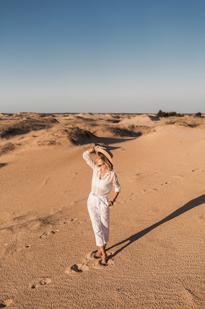 Stylish beautiful woman walking in desert sand in white outfit wearing straw hat on sunset