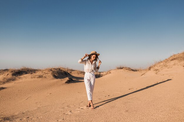 Stylish beautiful woman walking in desert sand in white outfit wearing straw hat on sunset