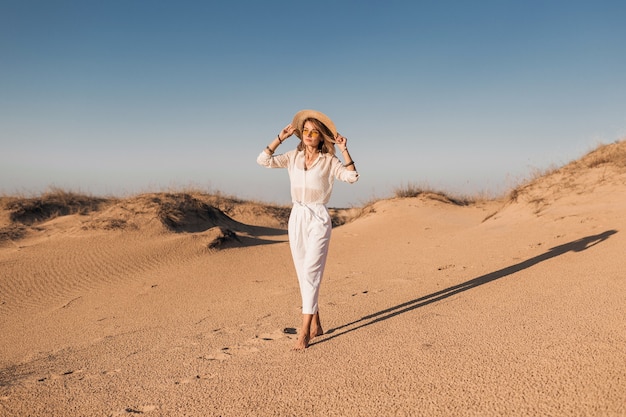 Stylish beautiful woman walking in desert sand in white outfit wearing straw hat on sunset