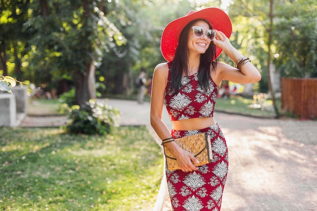 熱帯の衣装で公園を歩いているスタイリッシュな美しい女性。ストリートスタイルの夏のファッショントレンドの女性。わらのハンドバッグ、赤い帽子、サングラス、アクセサリーを身に着けています。休暇中に幸せな気分で笑っている女の子。