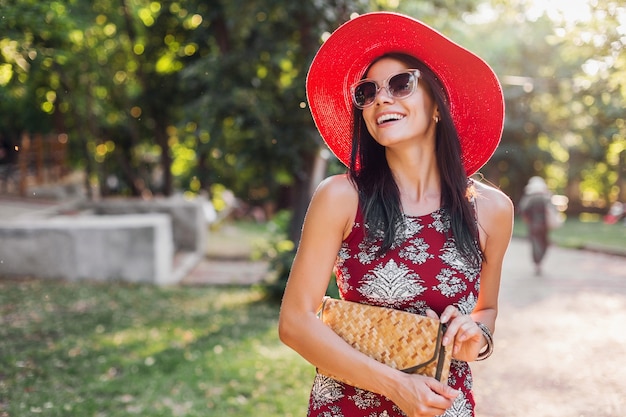 Stylish beautiful woman walking in park in tropical outfit. lady in street style summer fashion trend. wearing straw handbag, red hat, sunglasses, accessories. girl smiling in happy mood on vacation.