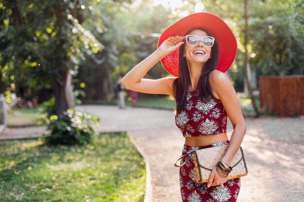 熱帯の衣装で公園を歩いているスタイリッシュな美しい女性。ストリートスタイルの夏のファッショントレンドの女性。わらのハンドバッグ、赤い帽子、サングラス、アクセサリーを身に着けています。休暇中に幸せな気分で笑っている女の子。