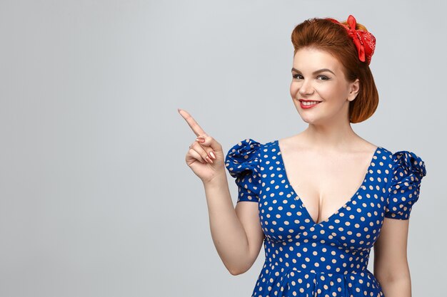 Stylish beautiful young red haired female wearing red lipstick and blue dotted dress smiling joyfully, indicating index finger at blank gray studio wall with copy space for your promotional content