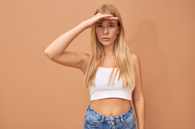 Stylish beautiful young woman in white top and blue jeans keeping palm on her forehead, protecting eyes from bright sunshine