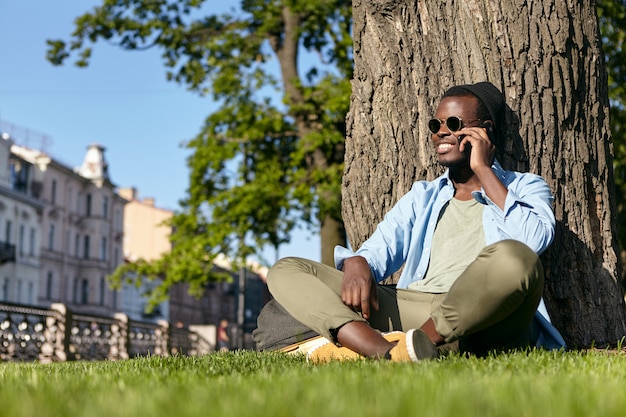 Stylish black hipster male wearing sunglasses, hat and shirt with trousers, sitting crossed legs at green lawn near big tree, having conversation on smart phone, enjoying beautiful weather and nature