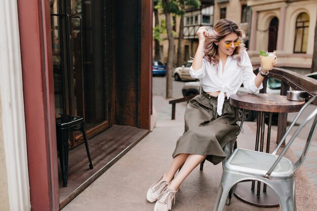 Stylish blissful girl in white shirt and long skirt drinks icy tea in outdoor cafe