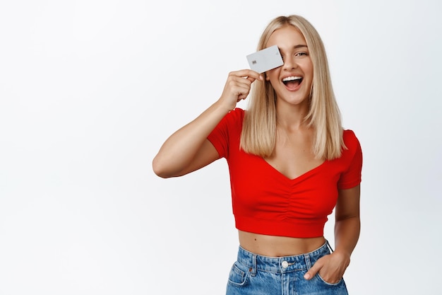 Stylish blond girl shows credit card near face and laughing concept of shopping and contactless payment standing over white background