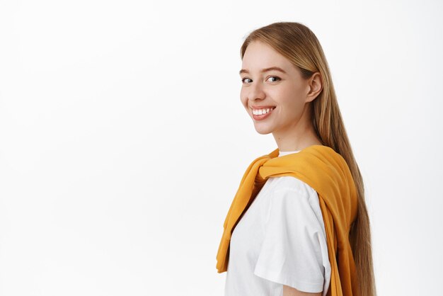 Бесплатное фото Стильная блондинка, стоящая в профиль, поворачивает голову в камеру с уверенной и самоуверенной улыбкой, стоя счастливой на белом фоне