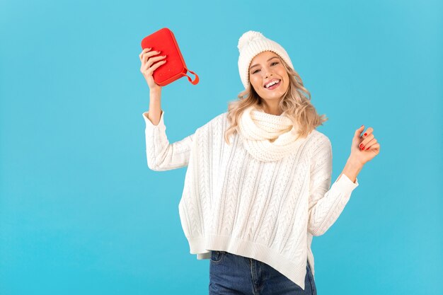 Stylish blond smiling beautiful young woman holding wireless speaker listening to music happy dancing wearing white sweater and knitted hat winter style fashion posing 