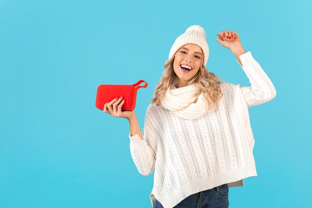 Stylish blond smiling beautiful young woman holding wireless speaker listening to music happy dancing wearing white sweater and knitted hat winter style fashion posing 