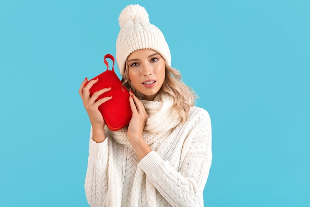 Стильная белокурая улыбающаяся красивая молодая женщина, держащая беспроводной динамик, слушает музыку, счастлива носить белый свитер и вязаную шапку, зимняя мода, позирует изолирована на синем фоне
