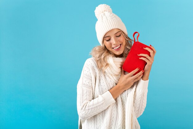 Stylish blond smiling beautiful young woman holding wireless speaker listening to music happy wearing white sweater and knitted hat winter style fashion posing 