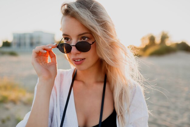 Стильная белокурая женщина в солнцезащитных очках наслаждается тропическим отдыхом на каникулах. Идеальная кожа. Естественная красота.