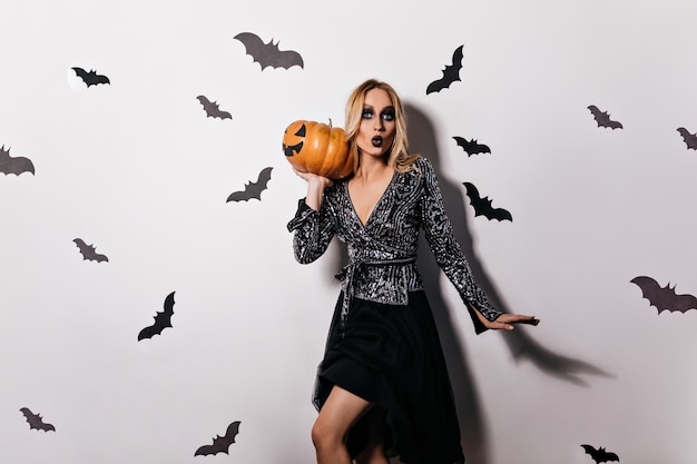 Стильная блондинка в черном платье позирует на вечеринке ведьм. Элегантная женская модель с темным макияжем, держащая большую тыкву на Хэллоуин.